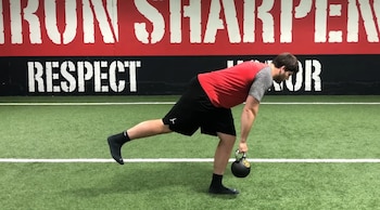Hombre de perfil realizando peso muerto unilateral con una kettlebell en la mano derecha, pierna izquierda extendida hacia atrás, sobre césped artificial