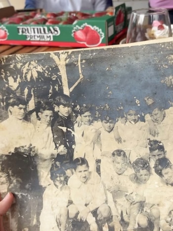 Una mano sostiene una fotografía antigua en blanco y negro de varios niños. Al fondo hay una caja de fruta sobre una mesa