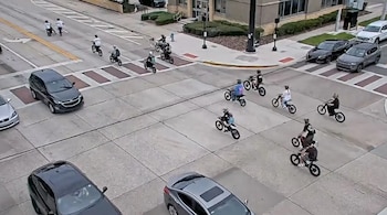 Trece personas en motocicletas eléctricas en una intersección urbana, junto a tres automóviles, señalización vial y edificios