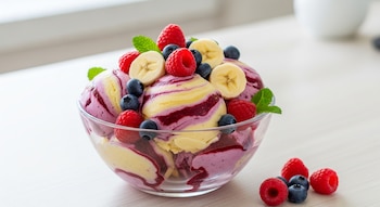 Tazón de cristal con helado morado y amarillo decorado con frambuesas, arándanos, rodajas de plátano y hojas de menta. Algunas bayas están sobre una mesa blanca.