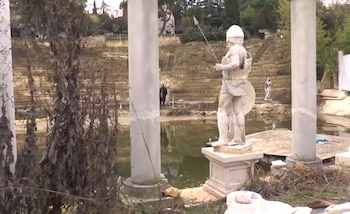 Estatua de piedra de un guerrero con casco y lanza, situada entre columnas y frente a un estanque y gradas de piedra