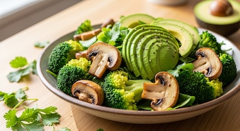 Ensalada con floretes de brócoli, champiñones en rodajas, y aguacate en abanico, espolvoreada con pimienta, sobre una mesa de madera clara. Medio aguacate al fondo.