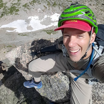 Selfie de Aron Ralston sonriendo, con casco verde, gorra roja, mochila y brazo protésico en un paisaje rocoso de montaña con nieve al fondo