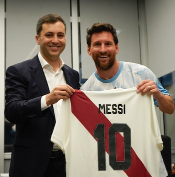 Dos hombres sonríen mientras sostienen una camiseta blanca de fútbol con una franja roja diagonal que dice MESSI 10