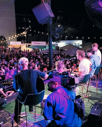 Músicos con guitarras sentados en un escenario al aire libre frente a una multitud, con un camarógrafo de espaldas en primer plano