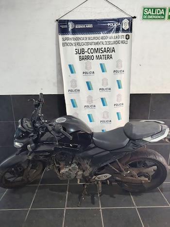 Motocicleta negra estacionada frente a un banner de la Policía de la Provincia de Buenos Aires y la Sub-Comisaría Barrio Matera. Se ve un cartel de "Salida de Emergencia"