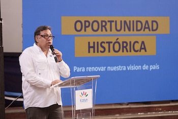 Hombre con camisa blanca y gafas habla en un micrófono desde un atril transparente. Detrás, una pantalla azul muestra "Oportunidad Histórica"