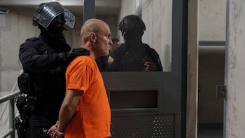 Un hombre con camiseta naranja y la cabeza rapada es custodiado por dos agentes con equipo táctico y casco