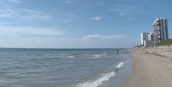 Una playa de arena con mar a la izquierda, edificios a la derecha y una avioneta a lo lejos que desciende sobre el agua