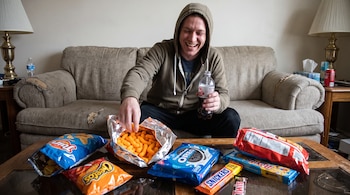 Hombre con sudadera en sofá sonríe. Toma snacks de una bolsa naranja abierta. En mesa hay paquetes de Cheetos, Tostitos, Oreo, Snickers, Twix y una botella de bebida.