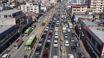 Vista aérea de una calle con múltiples carriles, vehículos de diversos tipos, autobuses, camiones, obras de construcción en el centro, y edificios a los lados.