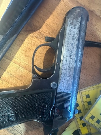 Una pistola semiautomática de metal sobre una mesa de madera, junto a un estuche negro y reglas amarillas de medición