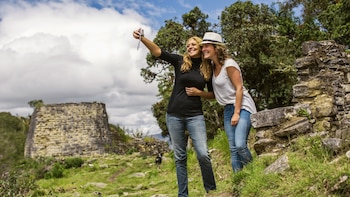 Dos mujeres sonríen mientras se toman una selfie en un sitio arqueológico de piedra con vegetación verde y un cielo nublado, un perro pequeño camina al fondo