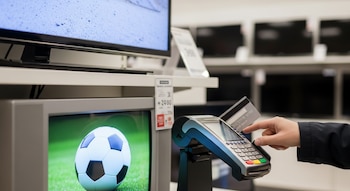 Una mano introduciendo una tarjeta de crédito en un terminal de punto de venta, junto a dos televisores, uno de ellos mostrando un balón de fútbol en la pantalla.