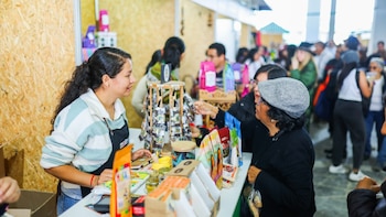 Una mujer con delantal y otra con gorra gris conversan en un puesto. Un mostrador blanco exhibe productos, paquetes y frascos. Personas al fondo