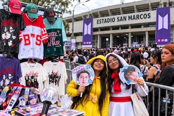 Dos jóvenes sonrientes sostienen carteles con rostros de músicos junto a un puesto con camisetas y gorras frente al Estadio San Marcos