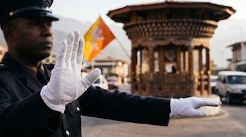 Un agente con uniforme y guantes blancos hace señas con las manos en una calle con vehículos, una glorieta tradicional y la bandera de Bután.