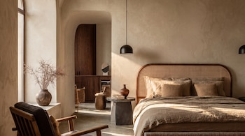 Dormitorio moderno con paredes de yeso color arena. Una cama con cabecero de ratán, ropa de cama de lino beige, mesita de piedra. Un sillón y jarrón con ramas cerca de una ventana.
