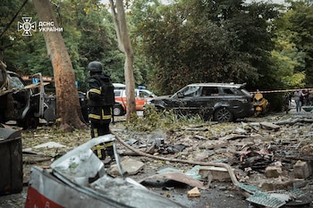 Hombre con casco y uniforme, vehículos siniestrados, camión, SUV negro, scooter amarillo, escombros en el suelo, árboles, vehículo de emergencia