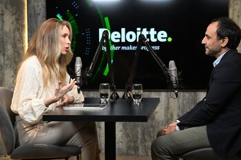 Una mujer y un hombre sentados ante una mesa negra con micrófonos y vasos de agua frente a una pantalla con el logotipo de Deloitte