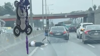 Una persona tendida en el asfalto, otra persona de pie, un automóvil oscuro, varios vehículos y un puente en una autopista con tráfico