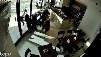 Vista desde una cámara de seguridad del interior de un café donde una persona vestida de negro rompe el cristal de la puerta de entrada
