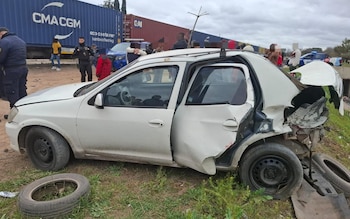 Automóvil blanco severamente dañado en la parte trasera y lateral, un neumático suelto en el suelo, contenedores de carga, personas y un vehículo policial