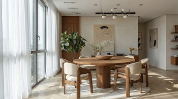 Comedor moderno con mesa redonda de madera clara y seis sillas tapizadas. Grandes ventanales con cortinas blancas transparentes y luz solar.