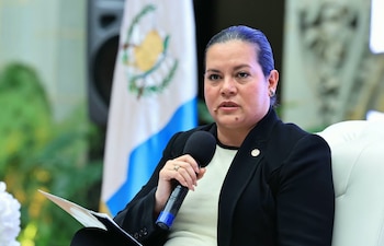 Una mujer vestida de saco negro sostiene un micrófono y papeles en la mano, ante la bandera de Guatemala