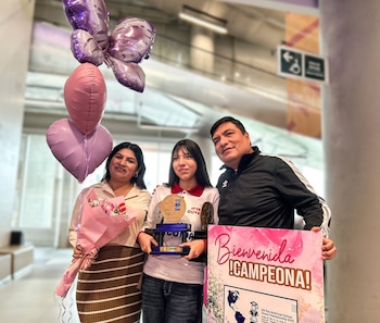 Aymé Maraví Cerón, una joven con camiseta blanca, sostiene dos trofeos. Una mujer con ramo de flores y un hombre la flanquean, junto a globos y un cartel de bienvenida