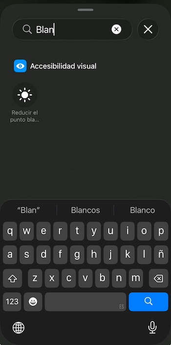 Una pantalla de teléfono móvil oscura con una barra de búsqueda que contiene la palabra "Blan". Debajo, se ven dos resultados: "Accesibilidad visual" y "Reducir el punto bla...". En la parte inferior, se visualiza un teclado virtual con sugerencias de palabras "Blan", "Blancos" y "Blanco"