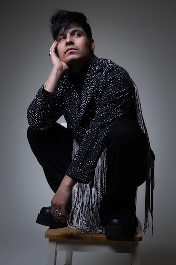 Louis David Horné, con chaqueta negra brillante y flecos plateados, está agachado sobre un taburete blanco. Mira hacia arriba con la mano en la barbilla y el fondo es gris liso