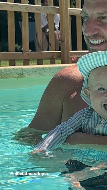 Un hombre sonríe mientras sostiene a un bebé que lleva un traje de baño y gorro a rayas en una piscina. Hay una valla de madera al fondo