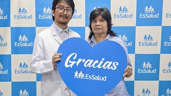 Un médico con bata blanca y una mujer de cabello oscuro sonríen mientras sostienen un cartel azul en forma de corazón con la palabra "Gracias EsSalud" en blanco