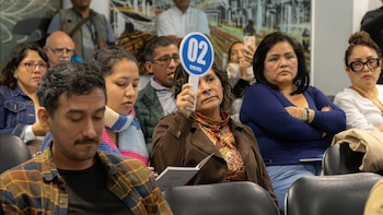 Varias personas sentadas en filas y una mujer en el centro que levanta una paleta azul con el número 02