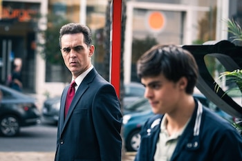 Un hombre de traje oscuro y corbata roja mira al frente; un joven de chaqueta oscura y camisa clara, desenfocado, se ve en una calle urbana