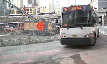 Un autobús de pasajeros en una calle junto a una cerca metálica, una señal naranja de obras y maquinaria de construcción al fondo