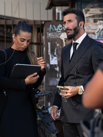 Una mujer y un hombre de pie, ambos con auriculares. La mujer mira una tablet, el hombre sostiene gafas de sol y un teléfono. Fondo urbano borroso