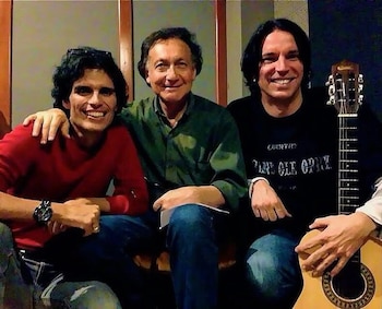 Tres hombres sonrientes sentados; el de la izquierda lleva una camiseta roja; el del centro, una camisa verde; el de la derecha, una camiseta negra y una guitarra acústica