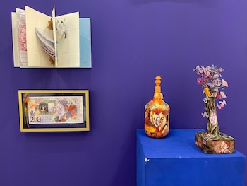 Un billete enmarcado, un libro desplegado en la pared, una botella pintada y una escultura de árbol sobre un podio ante un muro morado