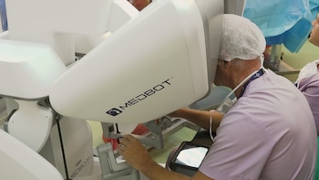 Un cirujano con gorro y mascarilla observa por el visor de un sistema de cirugía robótica Medbot mientras opera los mandos