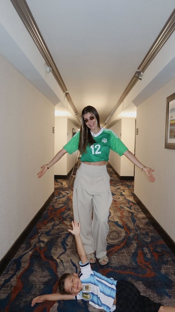 Mujer con camiseta verde y pantalón beige junto a un niño con camiseta de la Selección Argentina tumbado en el suelo de un pasillo de hotel