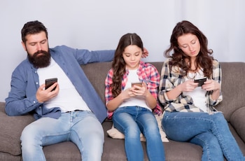Un hombre, una niña y una mujer sentados en un sofá gris, cada uno mirando y sosteniendo un teléfono móvil