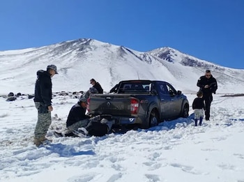 Varias personas y efectivos uniformados junto a una camioneta atascada en un camino nevado frente a montañas