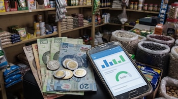 Billetes de peso mexicano y monedas apilados junto a un teléfono móvil con aplicación de banca digital y gráficos. Fondo de tienda con estanterías y productos.