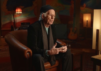 Keith Richards sentado en un sillón de cuero marrón, con un gorro negro y pañuelo estampado, en una habitación con instrumentos y arte