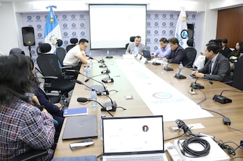 Sala de reunión con mesa rectangular, varias personas, micrófonos, botellas de agua, laptops, una pantalla de proyección, bandera de Guatemala y logo VALOR