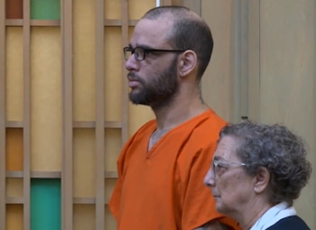 Un hombre con barba, anteojos y uniforme naranja de prisión permanece de pie junto a una mujer con lentes en una sala de tribunal
