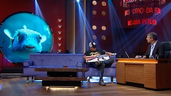 Hombre con gorra y keytar en sofá de estudio de TV iluminado. Detrás, pantalla con pez globo y presentador tras escritorio