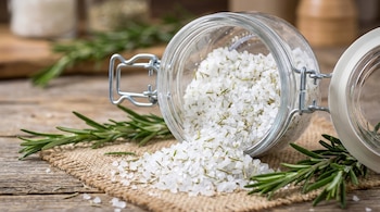 Un frasco de vidrio abierto está tumbado, derramando sal gruesa mezclada con hojas de romero sobre una tela de yute y una superficie de madera.